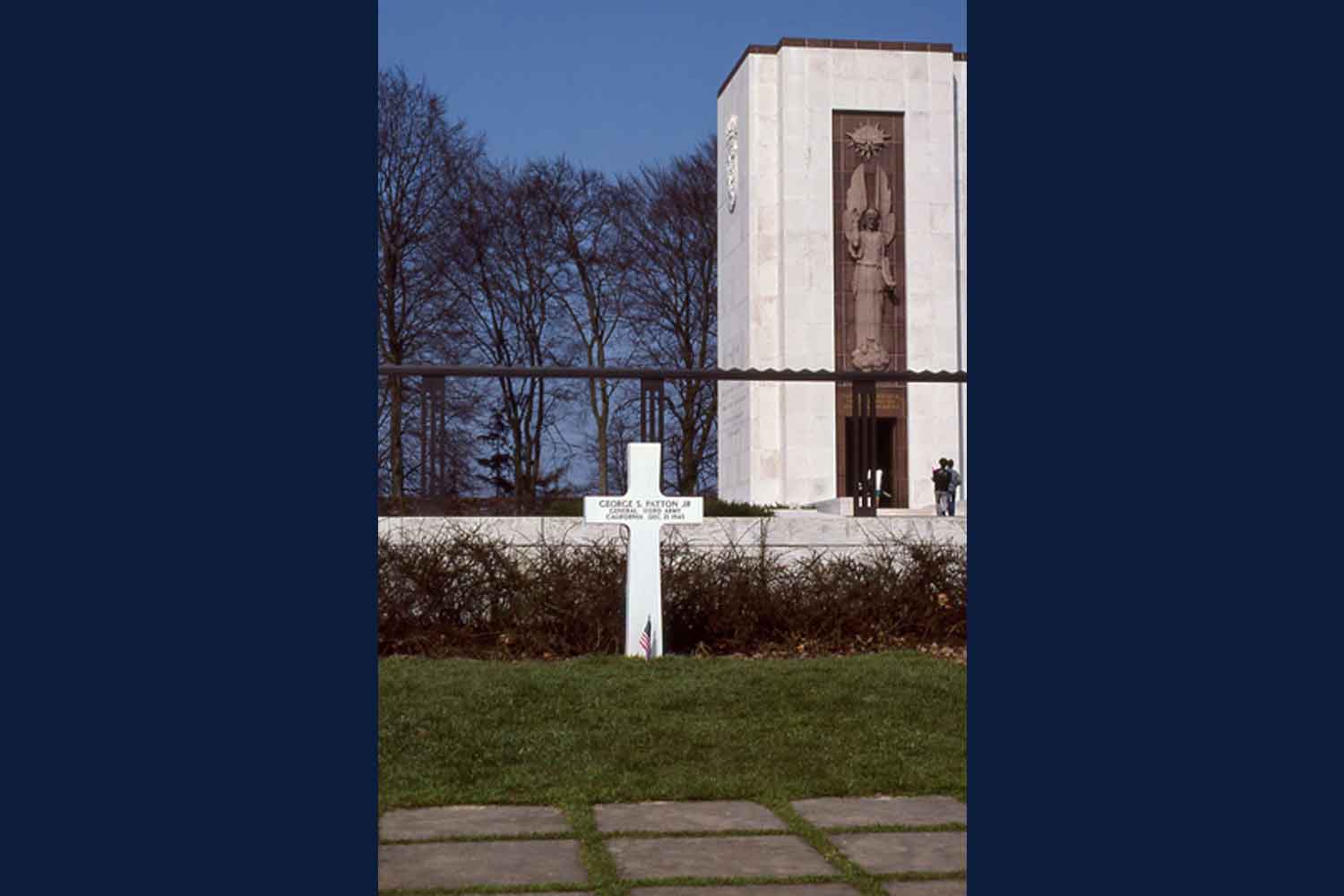 Grave site marked by a single cross with a large memorial in the background