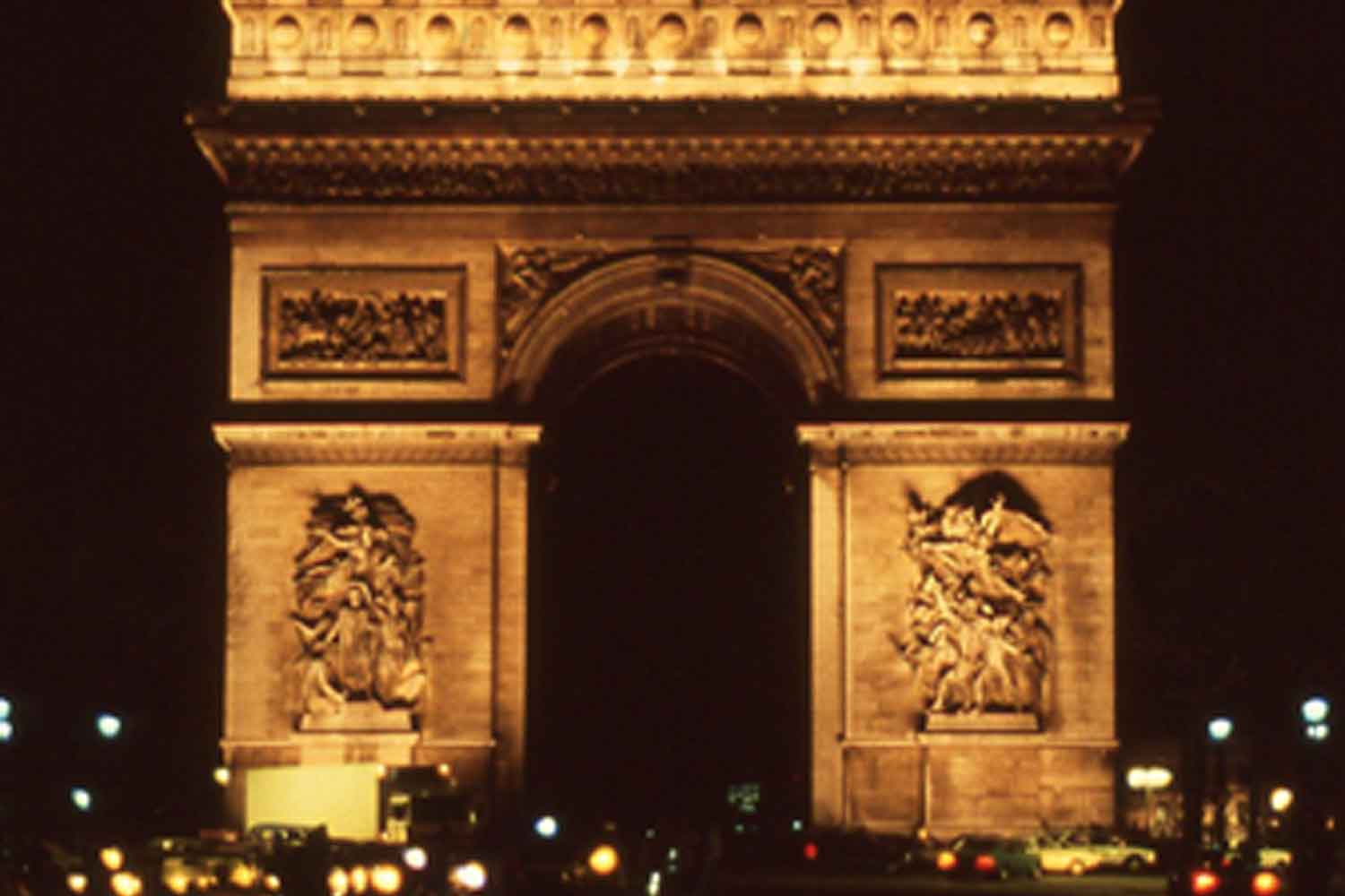 Photo of the Arc de Triomphe lit up at night. Large rectangular, decorated with reliefs and a large arched opening in the center through which traffic can drive.