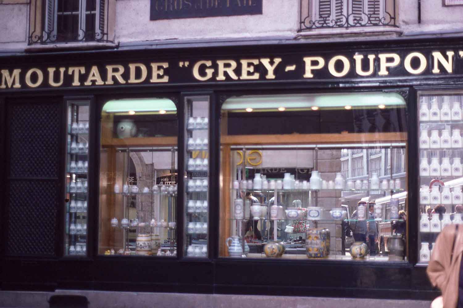 store front with signage of Moutarde Grey Poupon. Store window filled with white crocks holding Dijon Mustard