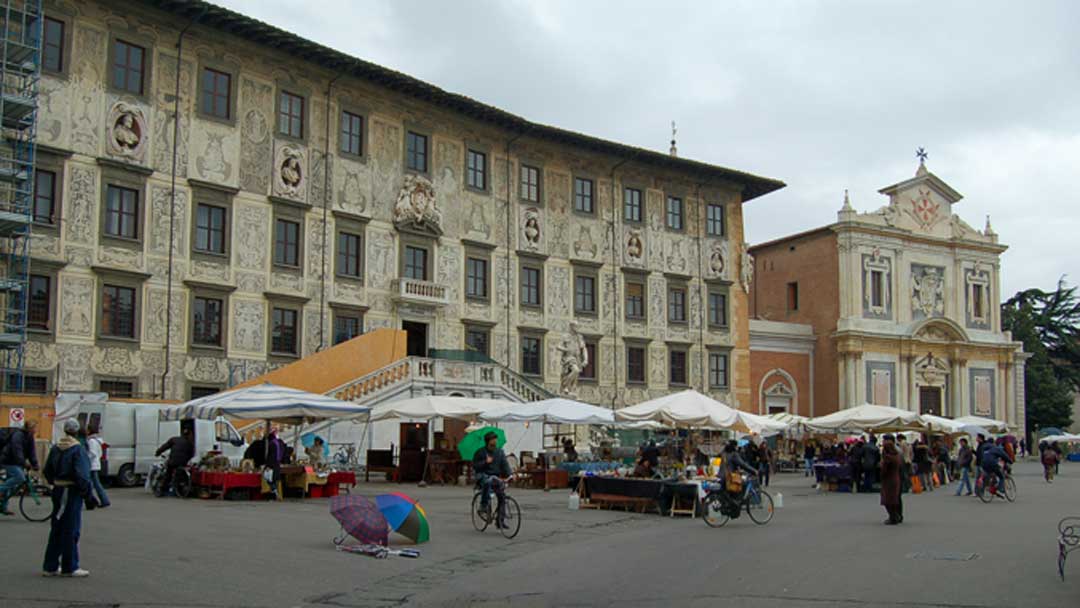 Long off white multi-story building with painted facade. A smaller church next to the building. Tents with people shopping in front of large building.