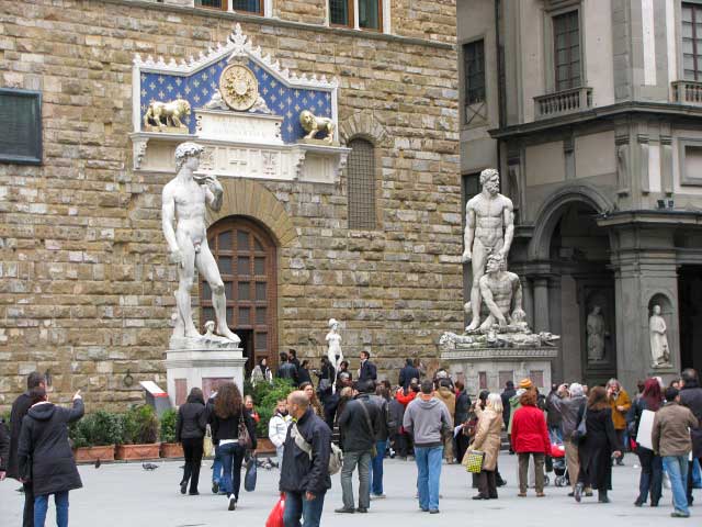 Tan stone building with a light blue sign over the door. Two large statues are outside of the building in the square. One is of a single man, the other is of two men.