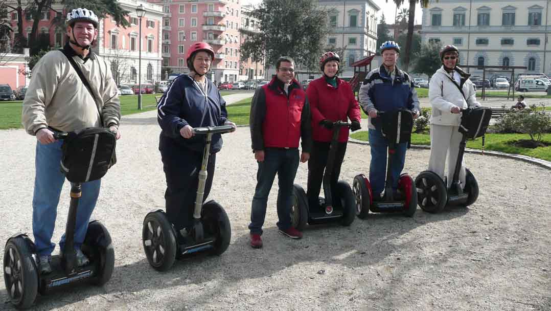 5 people standing on two wheeled gyroscopic vehicle, all in a row getting ready to go on a tour. One person in the middle is not on a vehicle.