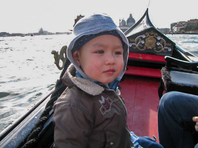 little boy about three years old with a had and hoodie pulled up over his head. He is wearing a jacket as well. He is riding in a gondola on the Grand Canal in Venice.