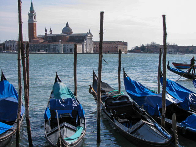 Photo of the bell tower and church in St. Marks Square, Venice taken from across the Grand Canal. In the foreground are five black gondolas covered with blue tarps. The gondolas are tied to poles protruding about 8 to 10 feet from the water's surface.