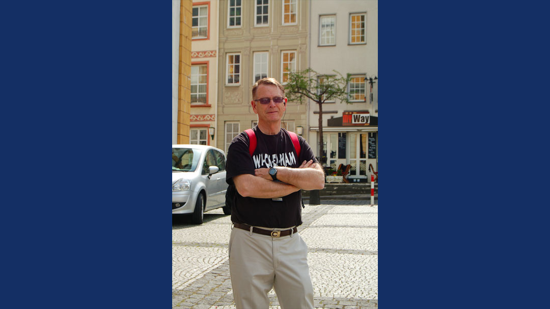 Man standing with arms crossed. he is wearing a black t-shirt with "wi-ck-er-ham" written on the front of it. He is standing outside in the sunshine, there are buildings behind him in the background.