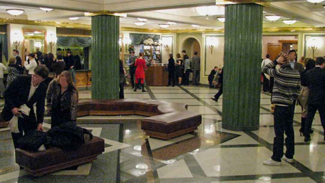 Lobby of Bolshoi Theater, large green marble pillars, floor of polished marble in white, green and red. a few benches in the center of the room. In background is coat check room