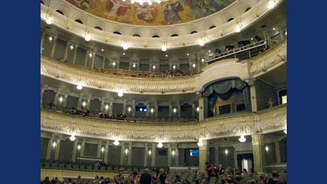 three tiers of seating in the New Stage of the Bolshoi theater painted domed ceiling. Large royal box with bluish gray curtains. Balconies of tiers are off white with raised decorations that resemble bunting with gold trim