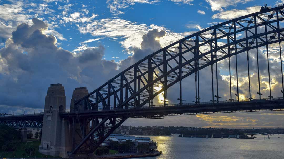 Pylons at the base of the arch of the Sydney Harbour Bridge. People climbing the bridge are visible on the arches. There are sailboats on the water in the background. There is a blue sky filled with large white clouds and the sun is setting behind the clouds casting a yellow glow on the water