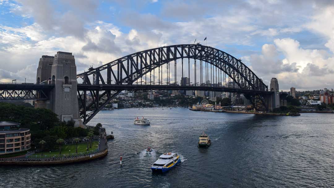More distant view showing the entire Sydney Harbour Bridge. Shows pylons of the bridge. Boats under the bridge and nearby. Blue sky with white and gray puffy clouds behind bridge.