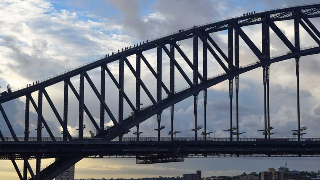 The arch of a suspension bridge, with people clearly walking on the the arch. Cloudy blue sky in the background.