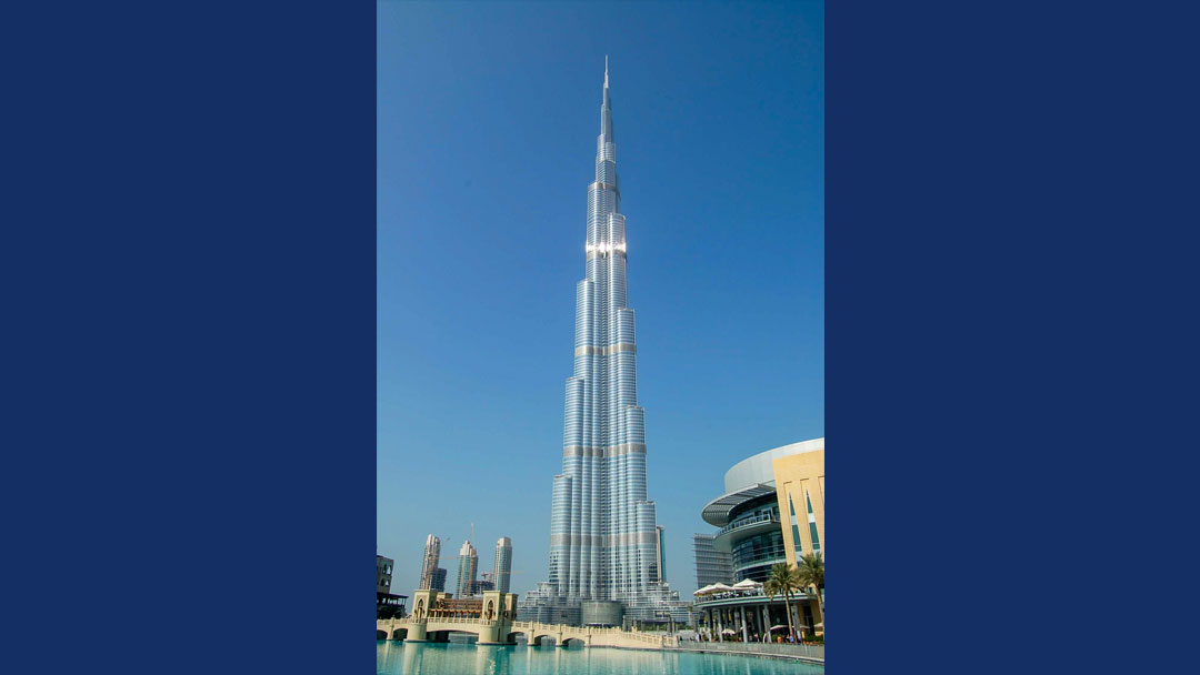 Tall thin tower. It looks like one tall cylinder with other cylinders added around it, with more added as it goes toward the ground. It is much wider at the bottom than the top. It appears silver in color with light glistening off it. There is a clear blue sky in the background. with smaller buildings around the base and a pool in the foreground.