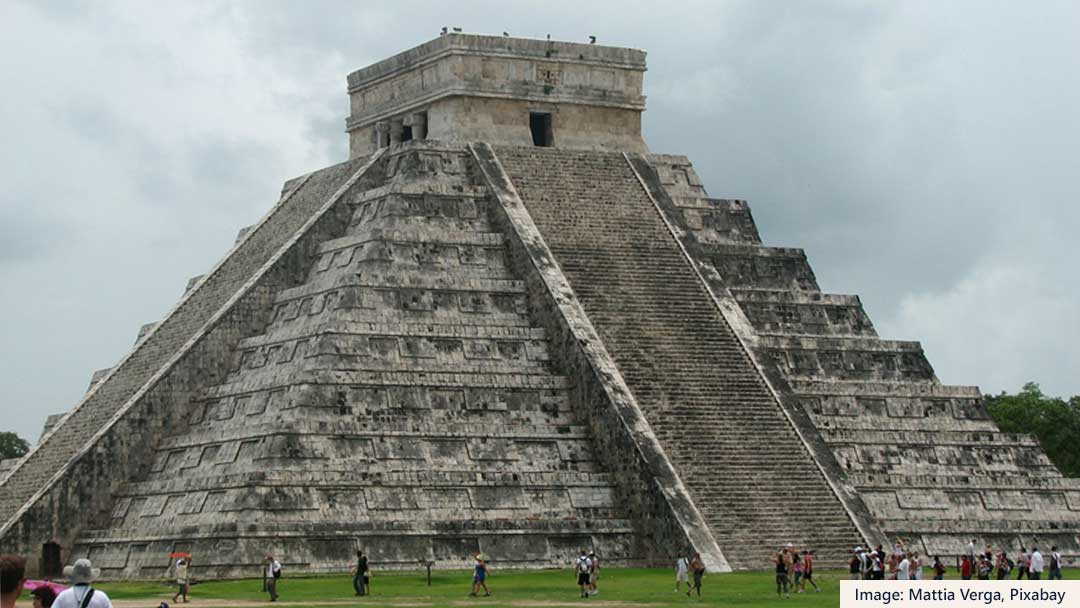 Large step pyramid, made of gray stones. Steps going up the center of the two visible sides. People walking in the grass in the foreground.