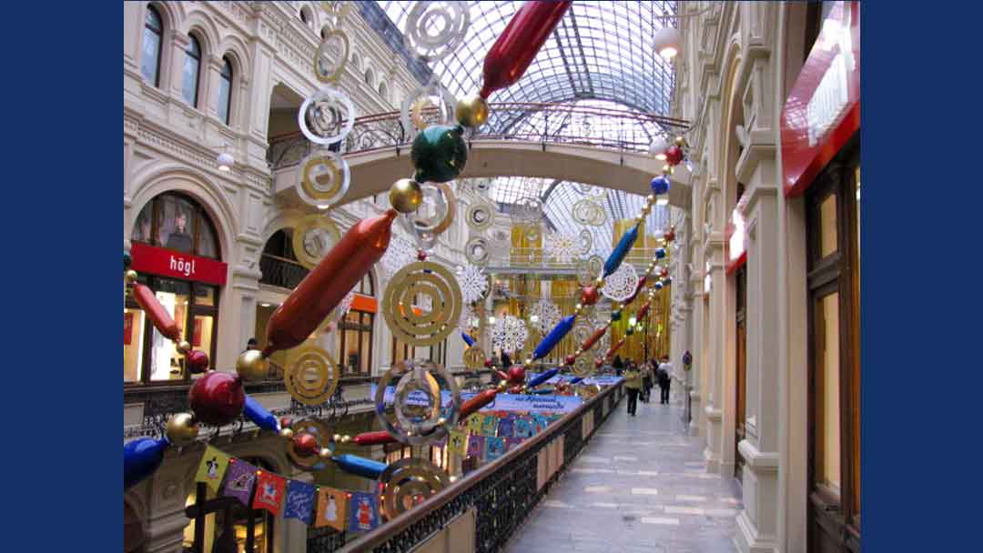 colorful garland draped from above second level hanging down to first level. Garland designed to look like beads in varying shapes in colors of blue, red, green and gold. Other hanging garland of silver and gold circle hung from ceiling. Ceiling is arched panels of glass.