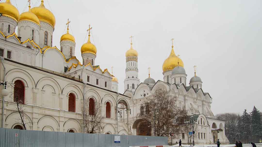 two large white brick buildings, one with three grey domed spires, and two gold domed spires, other church with 5 gold domed spires and gold trip on top level of roofline.