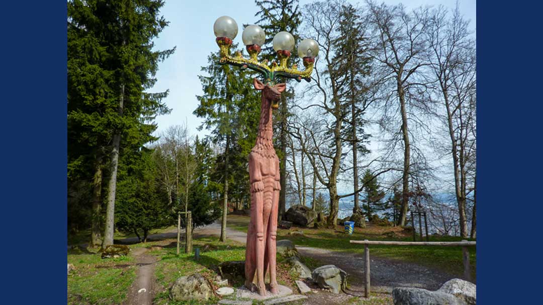 Whimsical light pole which looks like a caricature of a moose. it appears to be sitting, but has very long front legs and a rather long neck. There are antlers with large white round globes on top of the points, which are lights.