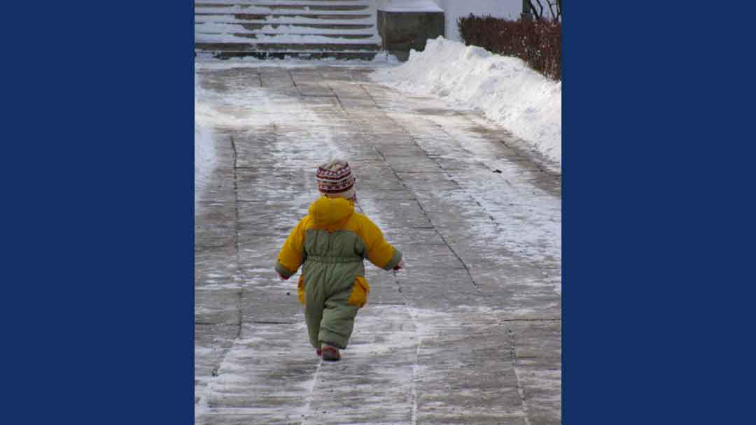 Child bundled in a greenish gray and orange snow suit wearing a knit hat walking along a brick walkway. the walkway has been cleared of snow and the snow is on piles on either side.