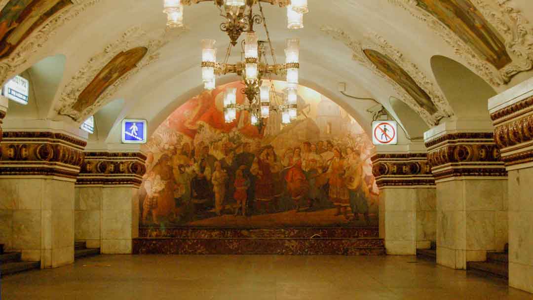 Arched hallway with a large mosaic depicting some time in Russian history, there is a marble bench in front of mosaic. There are columns holding up the arched ceiling. The square columns are covered in white marble. above the columns are mosaic pictures with stucco frames around each. There are three large chandeliers in the hall.