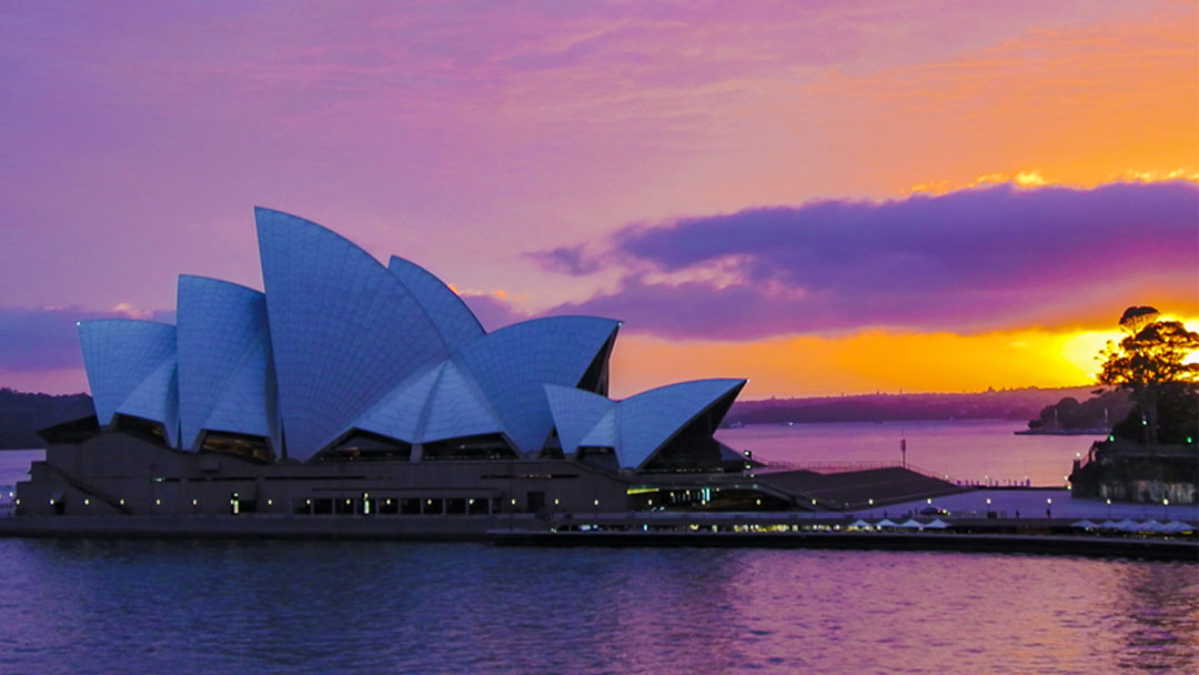 View of Sydney Opera House at Sunrise, sky tinted pink and purple from rising sun, clouds tinted purple as well. Large white Sydney Opera House comprised of arches covered in white tiles appearing blue in the early morning light, bright orange sun shining behind cloud in the back