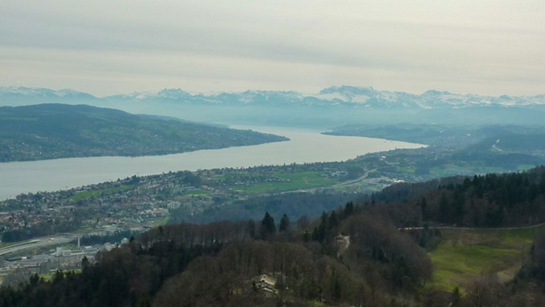 View from a mountain top of Alps mountains in the distance and a few buildings in the foreground with a long thin lake in the middle.