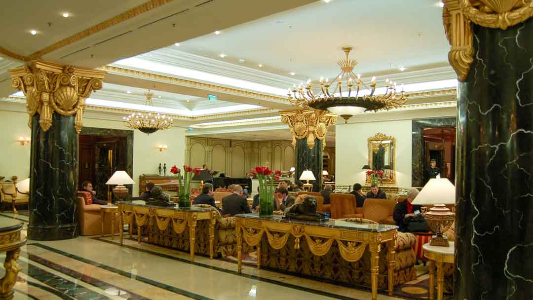 Interior hotel lobby filled with couches in dark brocade fabric with large easy chairs in a reddish gold fabric. Chandeliers hang from the ceiling. Two large black marble columns flank the room. On large tables behind sofas are vases with large red flowers in bloom. Floor is white marble with green and tan marble inlaid design. Tops of the columns and the tray ceiling are decorated in gold.