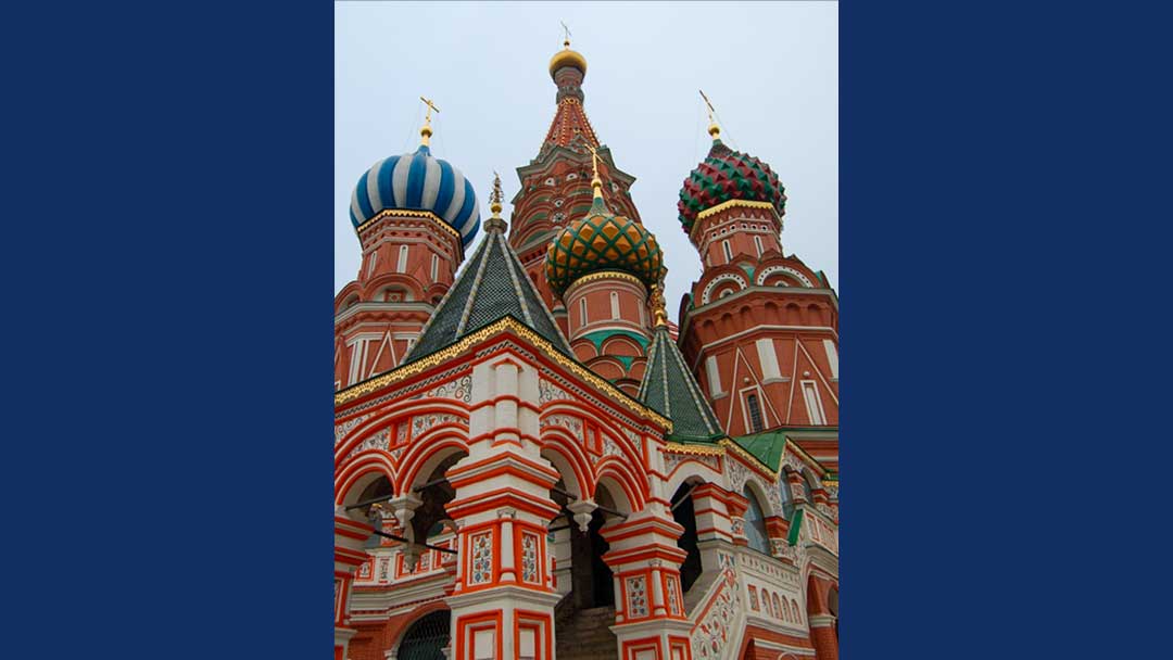Cathedral will multiple domes, each with different color and pattern. Gold crosses on top of Domes. One dome blue and white, one red and green, two are green and gold. Building is made of red brick.