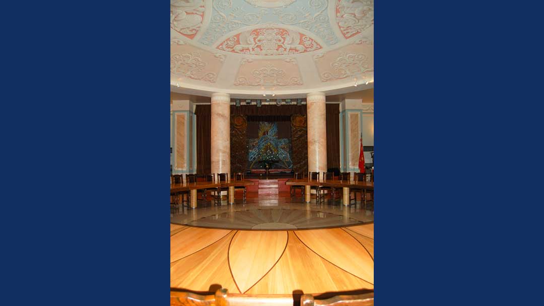 large round room with ornately painted ceiling with blues and peach tones. Peach marble columns support the domed ceiling. Lavish artwork on the back wall of the room. Large circular table made of wood with wood inlay