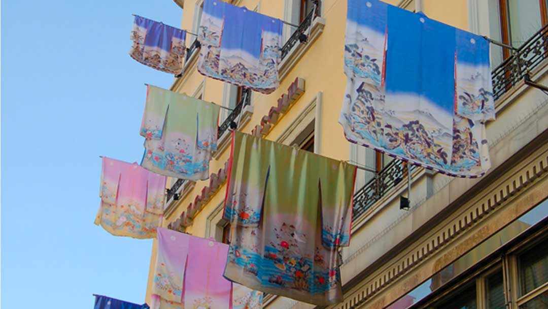 Bright colorful flags in the shape of kimonos hanging on poles from the side of a building, on three different levels of the building.