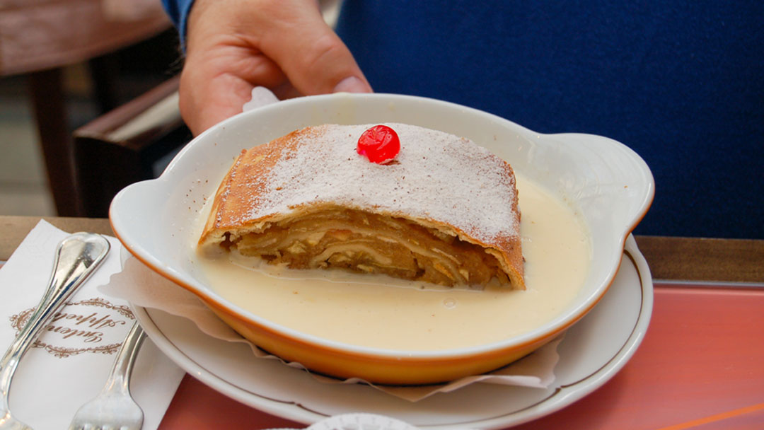 Slice of a large golden brown rolled pasty immersed in a white cream sauce, sprinkled with white powdered sugar and a bright red cherry on top.
