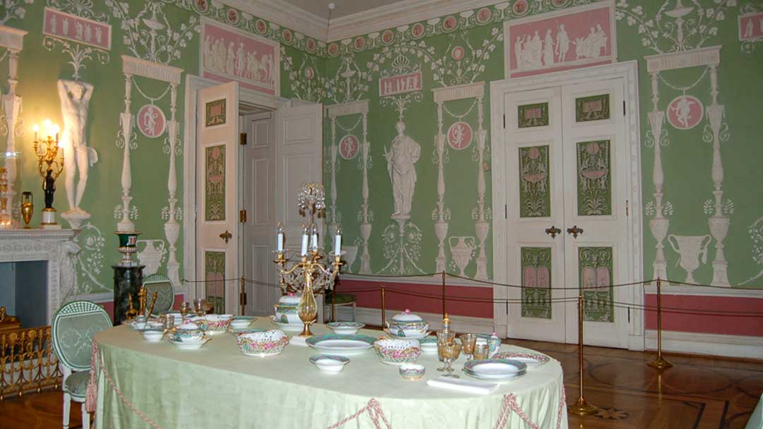 Dining room with walls painted green with friezes of people and urns in white. Pink inserts around the room also with friezes in white. Table is set with china.