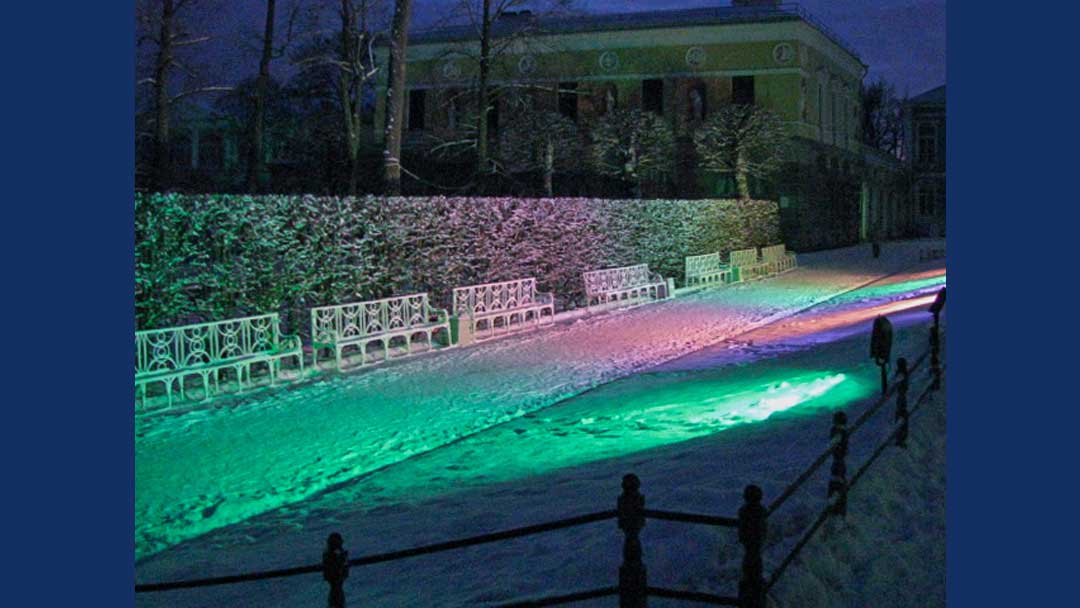 Multiple white benches in a row in front of leafless shrubbery. Snow on ground. Lit by multicolored spotlights. No people in photo but a few foot prints are in snow in front of benches.
