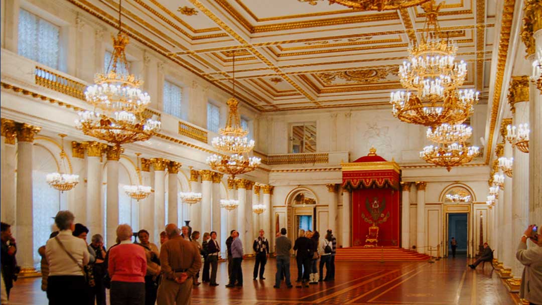 Large room with white marble pillars topped with gold leaves, room is all white finished with elaborate gold leaf trim. Ceiling is coffered with elaborate gold leaf trim. Floor is wood with elaborate inlay pattern. At end of room is a red canopy with gold trim under which sits a red chair with gold trim.