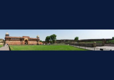 Panoramic view of red sandstone building with grass in front and tall wall to right of building.