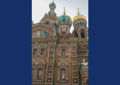 Exterior of church with multiple onion domes in a variety of colors. Onion domes are topped with gold crosses. In front of domes is a large mosaic. Cloudy sky in background.