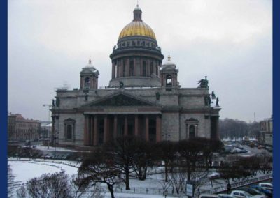 Large single domed church made of gray stone with a gold dome on the large cupola. The ground is snow covered and the sky is gray. There are leafless trees in the foreground.