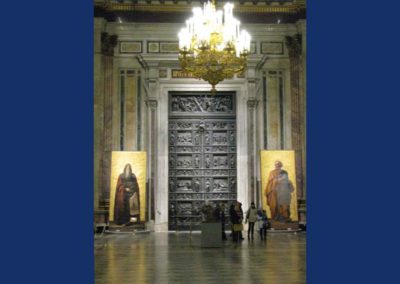 Large bronze door decorated with relief sculpture. Two large gold icons standing on either side of door. People in front of door demonstrating how large door and icons are.