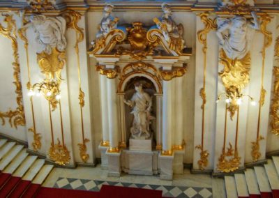 white marble double staircase coming down from both left and right. Wall is white marble with gold leaf decorations. In center platform where two stairs meet is a life size statue of a person, carved in white marble, in an alcove of white marble pillars topped with additional statues and gold leaf trim