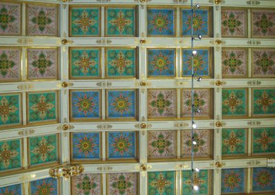 Ceiling of multiple squares. Squares are in pairs encased by white painted borders with gold trim. Individual squares are painted in colors of blue, green, pink. With different designs repeated within the squares.
