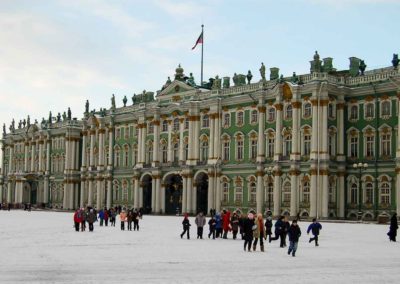 Exterior of building. Green walls. White pillars and window frames. Gold trim on pillars and window frames. Statues on tope of building. Russian flag flying over building. Children running in foreground. The ground is snow covered.