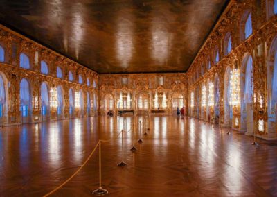 Very large room with numerous sets of windowed double doors, topped by arched windows reaching to the ceiling. The space between the windows and doors is covered in elaborate gold decorations. The floor is wood inlay. The ceiling is painted with a scene, and is very dark. There are sheer curtains over the windows and doors.