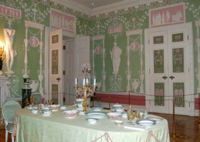 dining room with elaborately decorated walls. Walls are green with white relief decorations. Large white double doors decorated inserts are visible. There are several rectangular and circular areas framed in white with pink backgrounds. There are reliefs in white within the pink areas.
