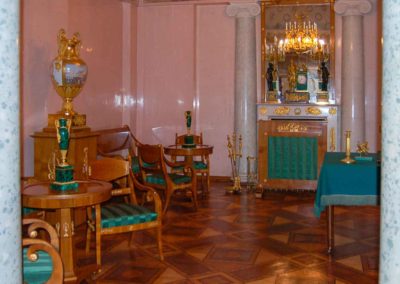 sitting room with pink walls, a fire place and multiple chairs, one large porcelain and gilt urn, two smaller jade and gold urns on separate tables. Wood inlay floor.