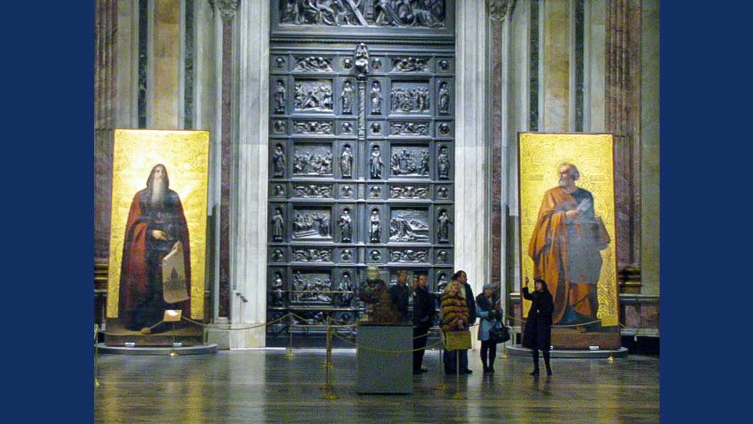 Large bronze door decorated with  relief sculpture. Two large gold icons standing on either side of door. People in front of door demonstrating how large door and icons are.