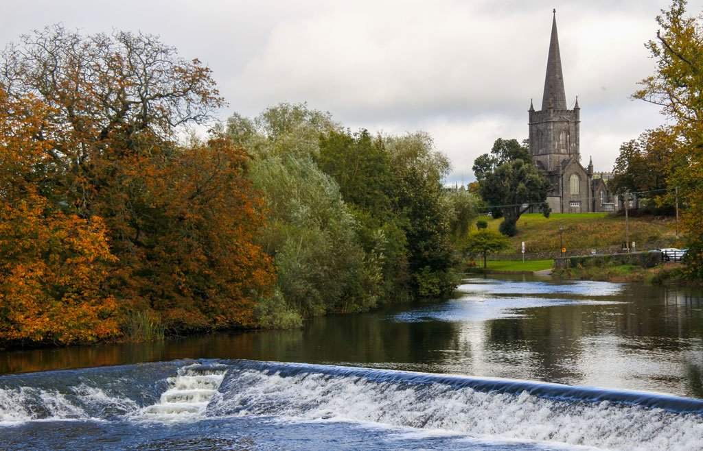 River flowing over a weir with fall colors on the trees and a church steeple in the background, Cahir, Ireland