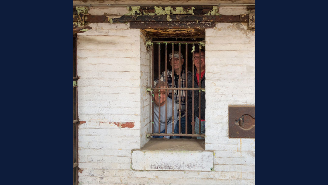Three men standing behind barred window within a cell at the Ohio State Reformatory. Men are tourists, dressed in modern day clothing and smiling at the camera. The Barred window is within a brick wall painted white, although the paint is peeling off the wall.
