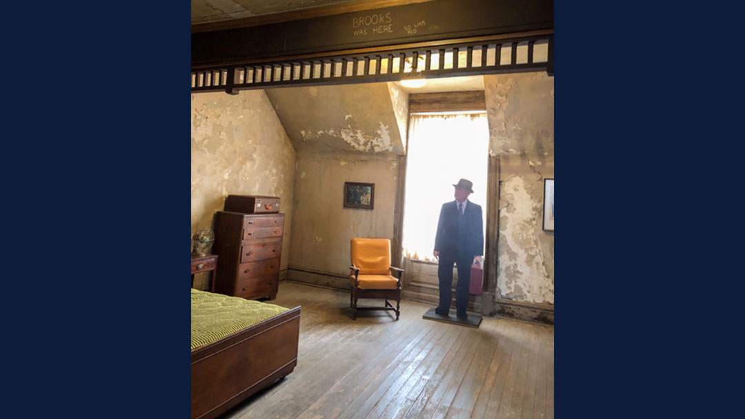 Set of a hotel room used by Brooks and Red in the movie The Shawshank Redemption. There is a single bed, a side table, a dresser with a suitcase on top, and one chair. There is a window covered by a curtain with sun shining through the fabric. There is a life sized cut out of the character of Brooks standing in front of the window. At the top there is a beam with the words "Brooks was here" and "So was Red" scratched in it. Paint is peeling off the walls.