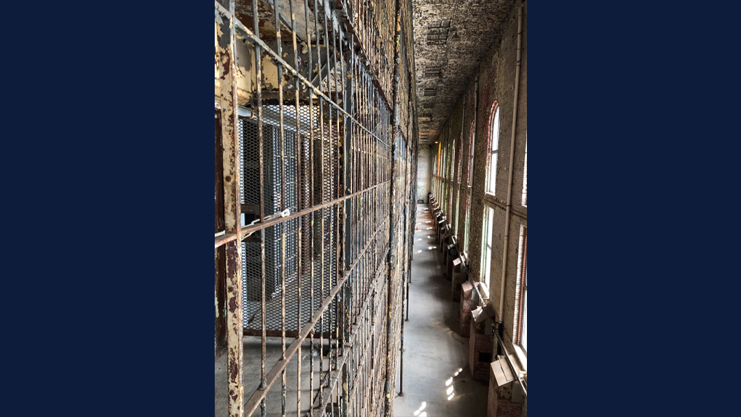 Large cage of prison steel prison cells showing multiple floors. Paint is peeling off ceiling, walls and bars. There are numerous large window allowing light into the room.